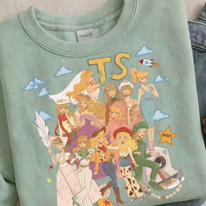 May include: A sage green crewneck sweatshirt featuring a graphic print of various animated characters dressed in diverse outfits, including a space suit and western attire. The text TS is printed in yellow above the characters, with a star labeled SHERIFY.