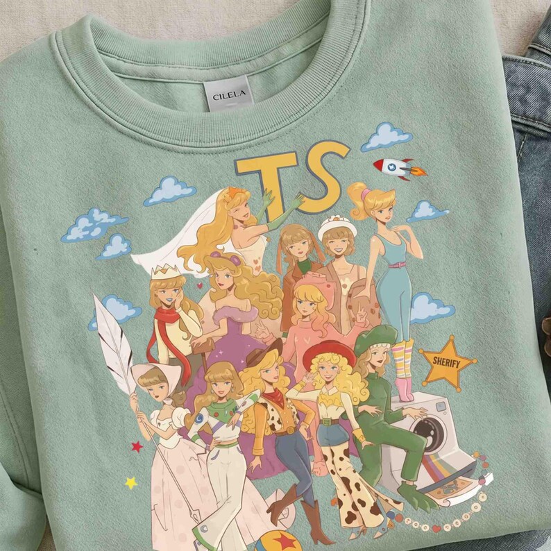 May include: A sage green crewneck sweatshirt featuring a graphic print of various animated characters dressed in diverse outfits, including a space suit and western attire. The text TS is printed in yellow above the characters, with a star labeled SHERIFY.