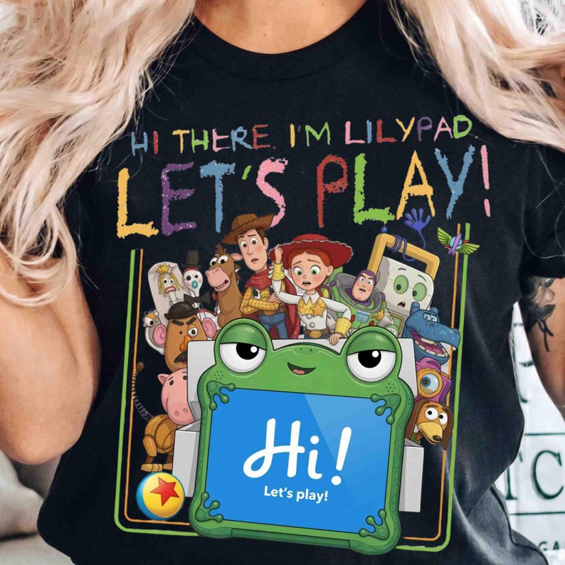 May include: A black t-shirt featuring a graphic print with the text "HI THERE, I'M LILYPAD. LET'S PLAY!" and "Hi! Let's play!". The design includes a green frog character holding a blue tablet surrounded by various animated toy characters.