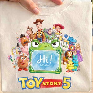 May include: A cream-colored t-shirt featuring a colorful graphic of Toy Story characters surrounding a green frog-shaped frame with the text "Hi! Let's play!" inside. Below the characters, the text "TOY STORY 5" is printed in large, bold letters.
