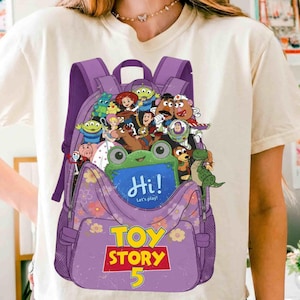 Pode incluir: Uma t-shirt cor de creme com um gr&aacute;fico de uma mochila roxa cheia de personagens de Toy Story. Um sapo verde segura uma placa azul que diz Hi! Let's play! O texto Toy Story 5 est&aacute; impresso em amarelo e vermelho abaixo da mochila.