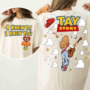 May include: A cream-colored t-shirt featuring a graphic print. The front displays the text I KNEW IT, I KNEW YOU TS STORY 5 with a heart. The back shows a person with long hair playing a guitar, a red cowboy hat, clouds, and TAY STORY text.