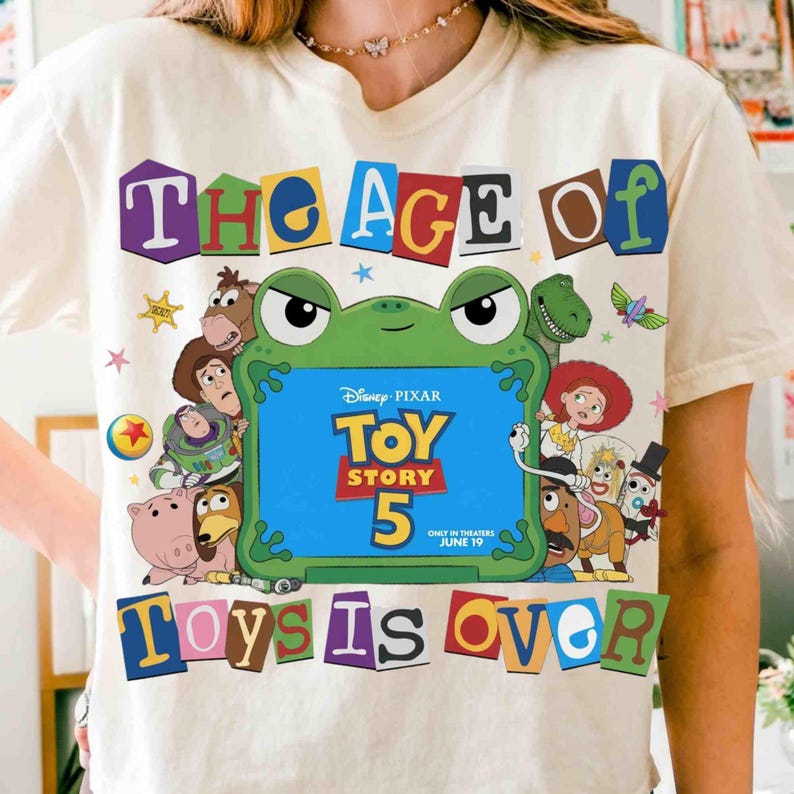 Pode incluir: Uma t-shirt de cor creme com uma moldura central verde em forma de sapo contendo o texto Disney Pixar Toy Story 5 Only In Theaters June 19. O design inclui v&aacute;rias personagens animadas a rodear a moldura com o texto The Age Of Toys Is Over.