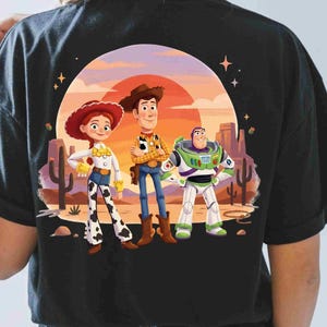 May include: A black t-shirt featuring a graphic print of Jessie, Woody, and Buzz Lightyear from Toy Story standing in a desert landscape with a large orange sun, cacti, and small stars against a sunset background with purple and orange hues.