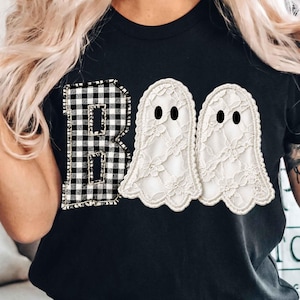 Może przedstawiać: Czarny t-shirt z grafiką przedstawiającą słowo BOO. Litera B wykonana jest w czarno-białą kratkę, natomiast dwie litery O przedstawione są jako białe duszki z koronki z czarnymi owalnymi oczami.