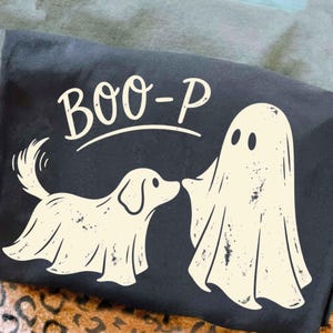 May include: A dark charcoal gray t-shirt featuring a cream-colored graphic of a dog and a ghost both draped in white sheets. Above the figures, the text BOO-P is printed in a cream-colored font with a curved line underneath the letters.