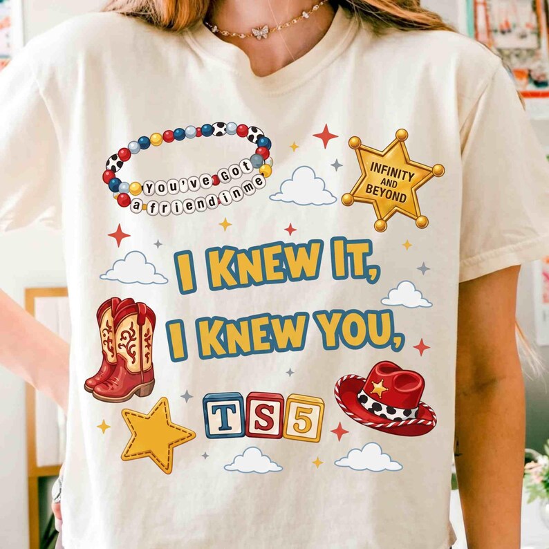 May include: A cream-colored t-shirt featuring a graphic print with the text "I KNEW IT, I KNEW YOU," a beaded bracelet with "YOU'VE GOT A FRIEND IN ME," a gold sheriff badge with "INFINITY AND BEYOND," cowboy boots, a red hat, and "TS5" blocks.