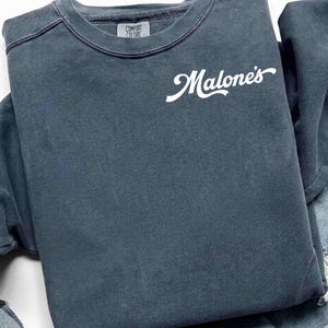 May include: A folded, slate blue sweatshirt with the word "Malone's" in white script across the front. The upper left corner features the "Comfort Colors" logo. The sweatshirt appears to be made of a soft, washed fabric.