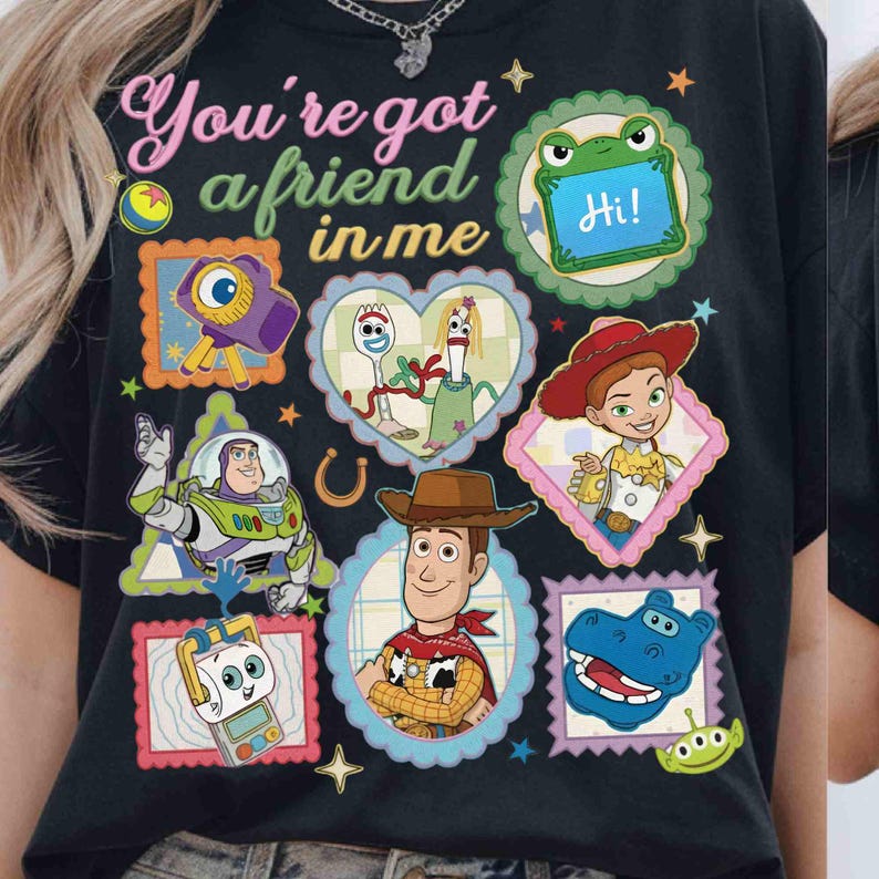 Puede incluir: Una camiseta negra con el texto "You're got a friend in me" rodeado de coloridas ilustraciones de personajes de Toy Story, incluyendo a Woody, Buzz Lightyear, Jessie, Forky y un alien&iacute;gena verde, dispuestos en varios marcos decorativos.