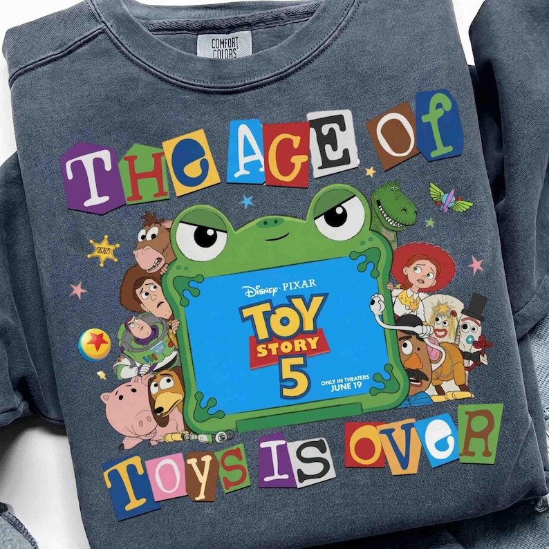 May include: A dark gray crewneck sweatshirt featuring a graphic print with the text THE AGE OF TOYS IS OVER. The design includes a green frog-shaped frame displaying the Disney Pixar Toy Story 5 logo and various animated character illustrations.