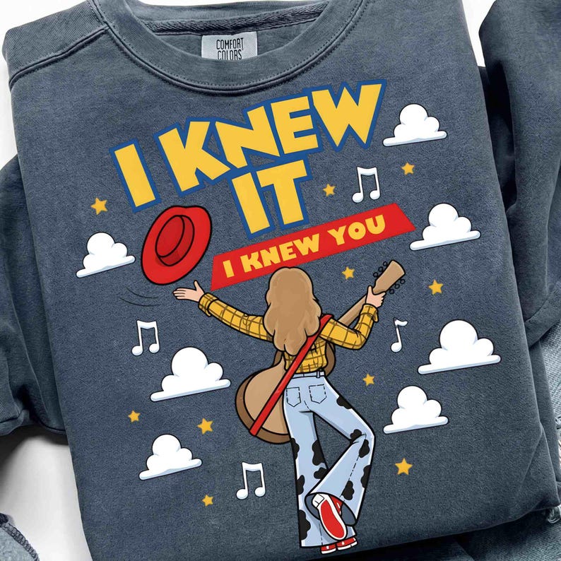 May include: A charcoal gray crewneck sweatshirt featuring a graphic of a person with long hair playing an acoustic guitar. The text reads I KNEW IT and I KNEW YOU, accompanied by illustrations of a red hat, white clouds, yellow stars, and music notes.