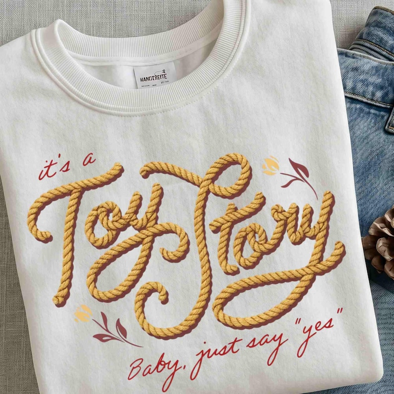 Puede incluir: Una sudadera blanca de cuello redondo con el texto "it's a Toy Story" escrito en una fuente amarilla estilo cuerda. Debajo del texto principal, la frase "Baby, just say "yes"" est&aacute; impresa en cursiva roja. Se aprecian peque&ntilde;os detalles florales rojos y amarillos.