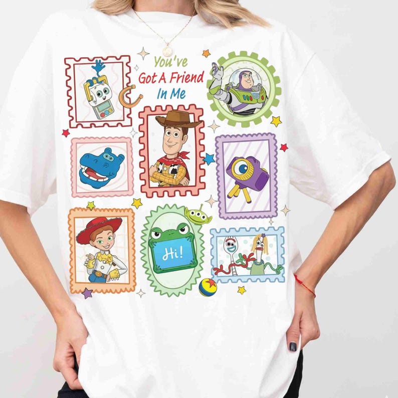 Puede incluir: Una camiseta blanca con un estampado gr&aacute;fico de varios personajes y objetos de Toy Story, incluidos Woody, Buzz Lightyear, Jessie y Forky, enmarcados en bordes de sellos postales coloridos con el texto You've Got A Friend In Me.