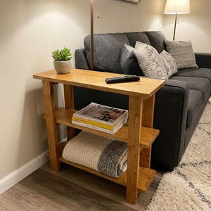 Peut inclure: Une console rectangulaire en bois avec une finition brun moyen, dot&eacute;e de trois &eacute;tag&egrave;res horizontales. La surface sup&eacute;rieure porte une petite plante succulente en pot et une t&eacute;l&eacute;commande noire. L'&eacute;tag&egrave;re centrale contient des magazines empil&eacute;s et l'&eacute;tag&egrave;re inf&eacute;rieure une couverture en tricot pli&eacute;e cr&egrave;me et grise.