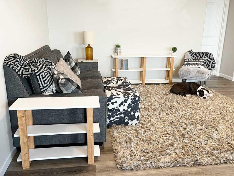 Peut inclure: Un canap&eacute; gris fonc&eacute; avec des coussins &agrave; motifs et un pouf &agrave; imprim&eacute; vache noir et blanc. Deux consoles blanches et en bois clair &agrave; trois &eacute;tag&egrave;res sont pr&eacute;sentes. Un tapis beige &agrave; poils longs recouvre le sol et un chien noir et blanc se repose.