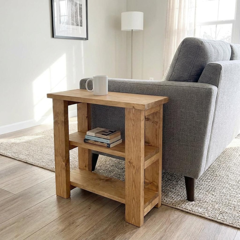 Peut inclure: Une table d'appoint rectangulaire en bois avec une finition brun clair naturel, comprenant un plateau plat, deux &eacute;tag&egrave;res de rangement ouvertes et quatre pieds robustes. Une tasse en c&eacute;ramique blanche est pos&eacute;e sur le dessus et deux livres sont empil&eacute;s.