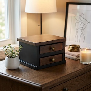 May include: A small, two-drawer wooden jewelry box with a dark brown top and drawer fronts, and a black body. The drawers have brass-colored knobs. The box sits on a wooden surface, next to a small potted plant and a lit candle.