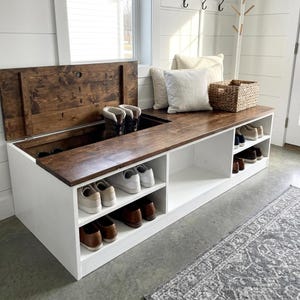Peut inclure: Un banc d'entrée en bois blanc avec un dessus en bois teinté brun foncé. Le banc dispose d'un compartiment de rangement à charnières sur le côté gauche, d'étagères ouvertes pour les chaussures en bas, et de deux coussins blancs et un panier.