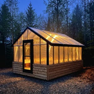 Op de afbeelding: Een houten kas met een helder dak en muren, van binnenuit verlicht. De constructie heeft een zwart deurkozijn en is omgeven door bomen. De buitenkant is gemaakt van lichtgekleurd hout en de binnenkant wordt verlicht met een warme gloed.