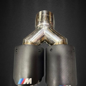 Könnte beinhalten: Doppelauspuffanlage mit Carbonfaser-Endrohren. Der Auspuff verfügt über ein poliertes Y-Rohr aus Edelstahl und zwei Carbonfaser-Endrohre mit dem "M"-Logo in Blau, Rot und Weiß. Die Endrohre haben ein strukturiertes Carbonfasergewebe.
