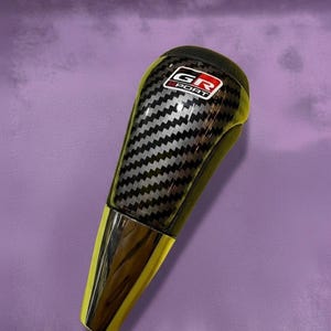 Peut inclure: Un pommeau de levier de vitesses en fibre de carbone noire et argentée avec une base chromée et des accents jaunes. Le pommeau porte le texte "GR SPORT" dans un rectangle blanc avec un bord rouge.