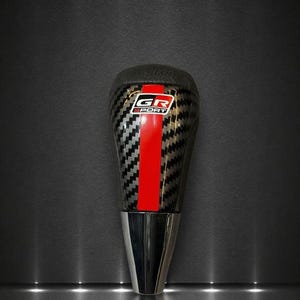 May include: A carbon fiber shift knob with a red stripe down the center. The top of the knob is black leather, and the base is chrome. The text "GR SPORT" is visible on the top of the knob.