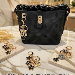 May include: Black bucket bag with embossed floral pattern and a chain strap. The bag features a gold-tone clasp and a charm with the letter "M" and black flower accents. The text "MAKE IT YOURS. WEAR YOUR INITIAL." is at the bottom.