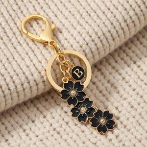 May include: Gold-tone keychain with a lobster clasp, a round ring, and a charm with the letter "B". The keychain features three black flower charms with gold details. The background is a cream-colored knitted fabric.