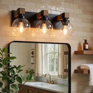 May include: A three-light vanity fixture featuring a matte black metal backplate, wooden accents, and three clear glass globe shades. Each globe houses a visible filament light bulb, providing warm illumination above a bathroom mirror and sink.