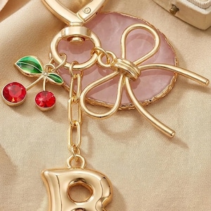 May include: Gold-toned keychain with a heart-shaped clasp. Features a pink stone accent, a bow, a chain, and a charm with the letter "B". Includes a cherry charm with red gems and green leaves. The keychain is on a beige background.