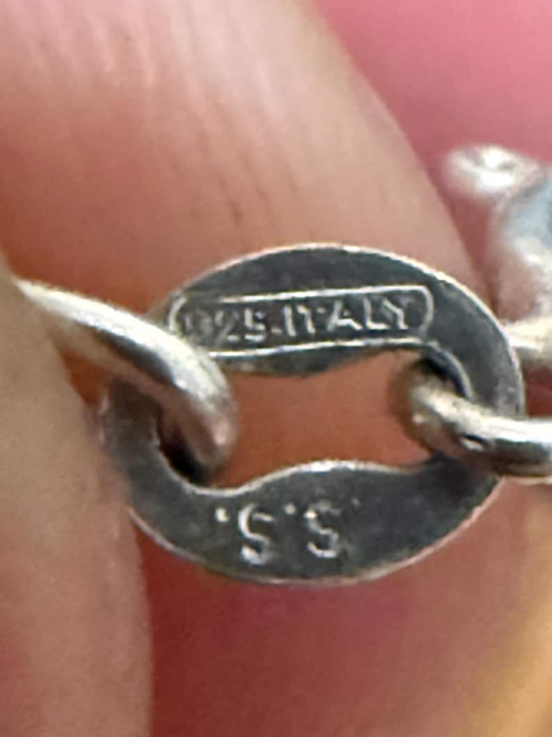 May include: Close-up of a silver jewelry clasp. The clasp is oval-shaped with the markings "925-ITALY" and "S.S." stamped on it. The clasp is attached to a silver chain, with a blurred pink background.