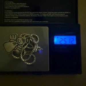 May include: A silver-colored necklace with a pendant featuring a cartoon angel design, resting on a digital scale. The scale displays a weight of 25.12, likely in grams or ounces. The necklace is on a square metal surface.