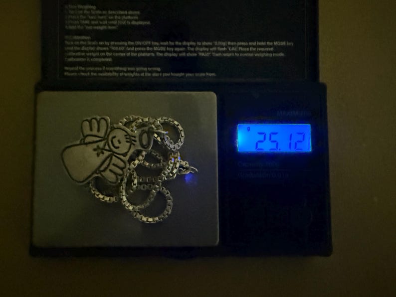 May include: A silver-colored necklace with a pendant featuring a cartoon angel design, resting on a digital scale. The scale displays a weight of 25.12, likely in grams or ounces. The necklace is on a square metal surface.