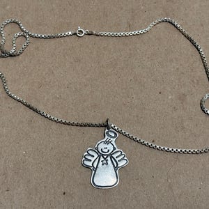 May include: Silver-toned necklace featuring a pendant of a smiling angel with a halo and outstretched wings. The chain is a delicate box chain style. The pendant is approximately 1 inch in height.