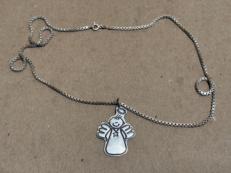 May include: Silver-toned necklace featuring a pendant of a smiling angel with a halo and outstretched wings. The chain is a delicate box chain style. The pendant is approximately 1 inch in height.