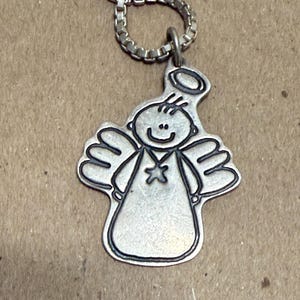 May include: A silver-colored pendant necklace featuring a cartoon angel design. The angel has a halo, wings, and a star on its chest. The necklace chain is a box link style. The pendant is approximately 1 inch in height.