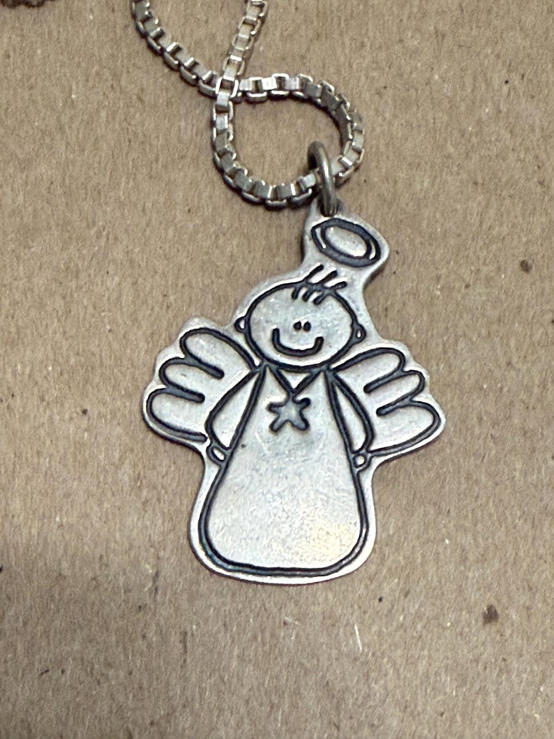 May include: A silver-colored pendant necklace featuring a cartoon angel design. The angel has a halo, wings, and a star on its chest. The necklace chain is a box link style. The pendant is approximately 1 inch in height.