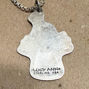 May include: A silver-colored pendant necklace with a unique, irregular shape. The pendant is attached to a silver chain. The pendant has the text "LUCY ANN STERLING USA" stamped on it.