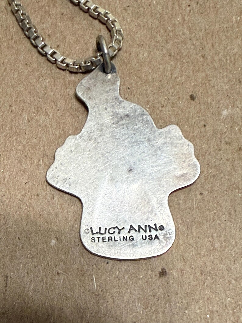 May include: A silver-colored pendant necklace with a unique, irregular shape. The pendant is attached to a silver chain. The pendant has the text "LUCY ANN STERLING USA" stamped on it.