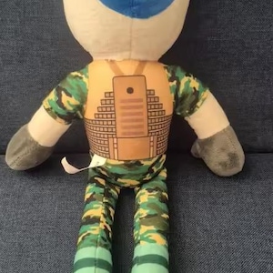 May include: Plush doll with a blue head, white face, and gray arms. The doll wears a camouflage shirt and pants, with a brown vest featuring a rectangular design. The feet are yellow.