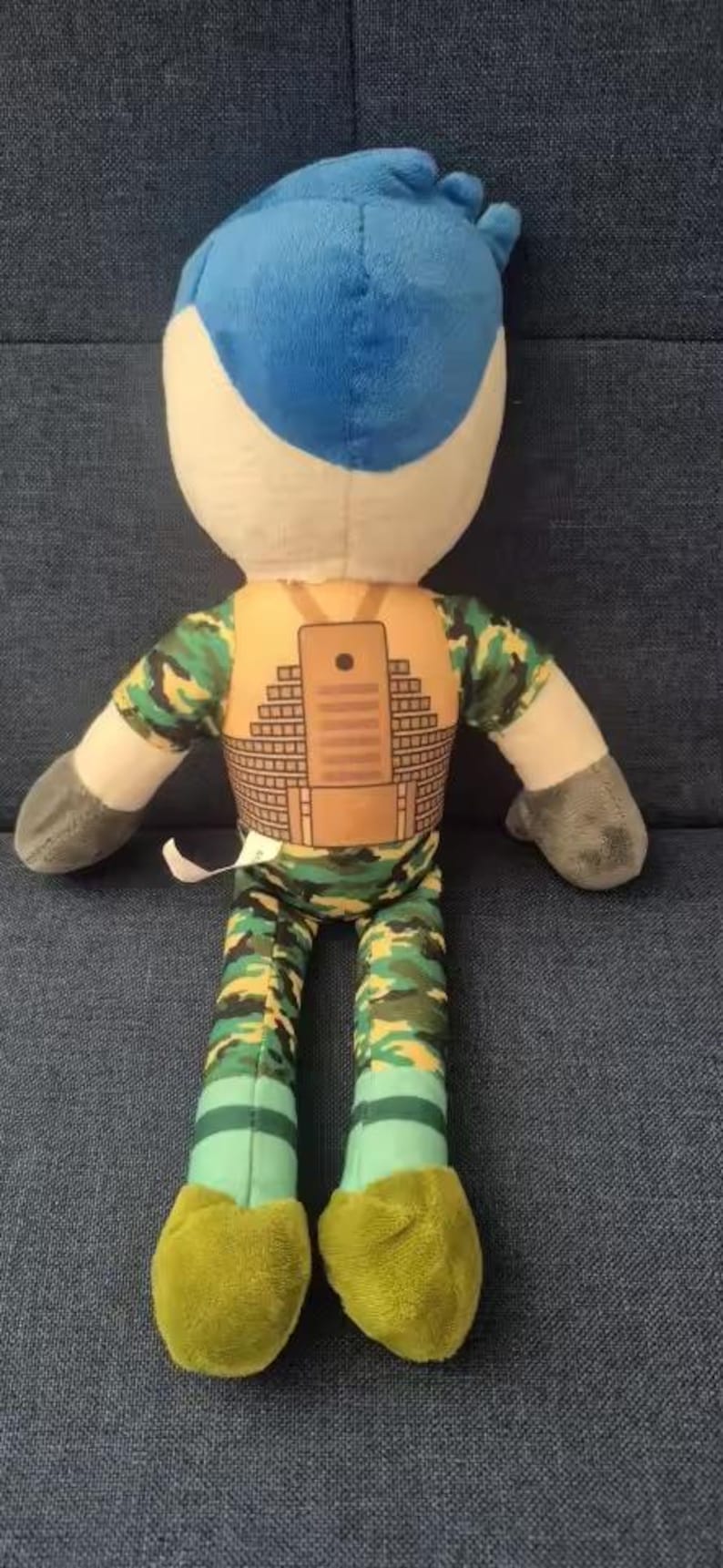 May include: Plush doll with a blue head, white face, and gray arms. The doll wears a camouflage shirt and pants, with a brown vest featuring a rectangular design. The feet are yellow.