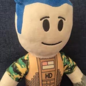May include: A plush doll with a blue hairstyle and a neutral expression. The doll wears a camouflage patterned outfit with an orange vest. The vest has the letters "HD" printed on it. The doll's arms and legs are also visible.