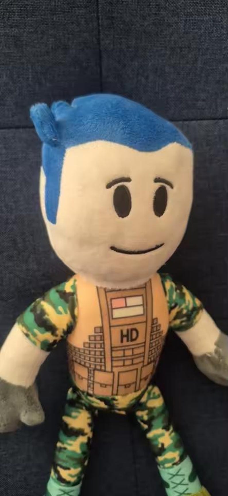 May include: A plush doll with a blue hairstyle and a neutral expression. The doll wears a camouflage patterned outfit with an orange vest. The vest has the letters "HD" printed on it. The doll's arms and legs are also visible.