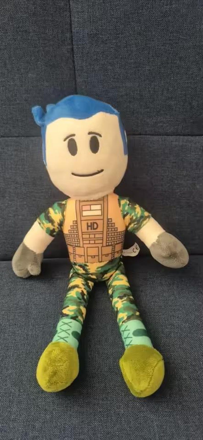 May include: A plush doll with a blue hair, a smiling face, and gray hands. The doll wears a camouflage vest and pants. The doll's feet are yellow.