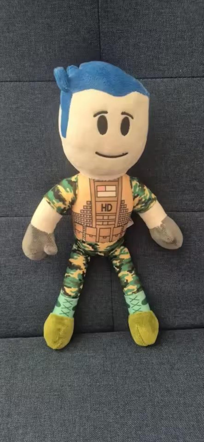 May include: A plush doll with a light skin tone, blue hair, and a smiling face. The doll wears a camouflage-patterned outfit with the letters "HD" on the chest. The doll's hands are gray, and the feet are yellow.