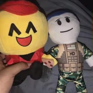 May include: Two plush dolls are held in a hand. One is yellow with a red hat and a smiling face. The other is a white doll with a blue cap, wearing a camouflage outfit with "HD" printed on the chest.