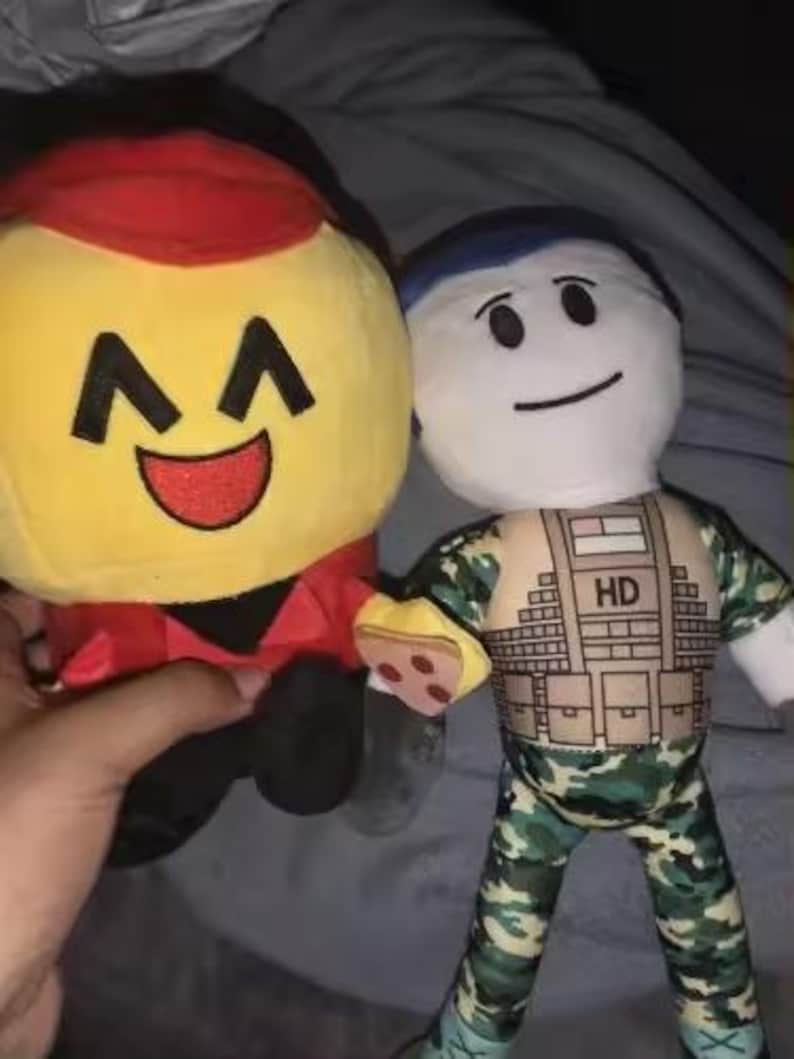 May include: Two plush dolls are held in a hand. One is yellow with a red hat and a smiling face. The other is a white doll with a blue cap, wearing a camouflage outfit with "HD" printed on the chest.