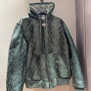 May include: A green and black snakeskin-patterned hooded jacket. It has a full-length zip, a drawstring hood, and a heart-shaped pendant. The jacket is displayed on a hanger.