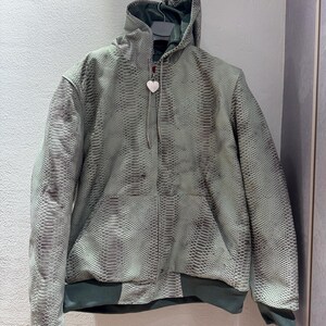 Puede incluir: Chaqueta con capucha con estampado de piel de serpiente en tonos grises y verdes. La chaqueta tiene cremallera frontal, cordón y un dije en forma de corazón. Los puños y el dobladillo son de color verde oscuro.