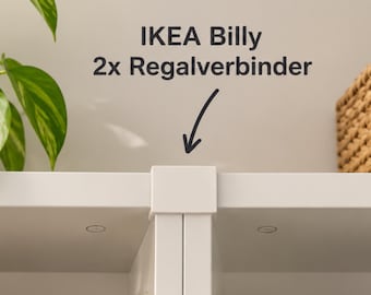 May include: A white plastic IKEA Billy 2x Regalverbinder shelf connector piece installed on the top edge of two adjacent white bookshelves. The text IKEA Billy 2x Regalverbinder is printed above the connector with a black arrow pointing down.